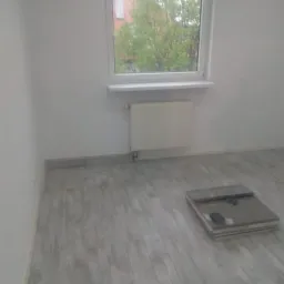 Szare panele podłogowe ułożone w pokoju z białymi ścianami, widoczne okno z widokiem na drzewo i grzejnik, paczka paneli na podłodze.