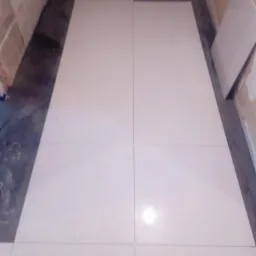 Układanie jasnych, błyszczących płytek podłogowych w trakcie prac wykończeniowych, widoczne niedokończone fragmenty podłoża i pionowo ustawiona płytka przy ścianie.