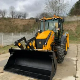 Żółto-czarna koparko-ładowarka JCB z opuszczoną łyżką na betonowej nawierzchni; w tle betonowe ogrodzenie i drzewa bez liści pod pochmurnym niebem.