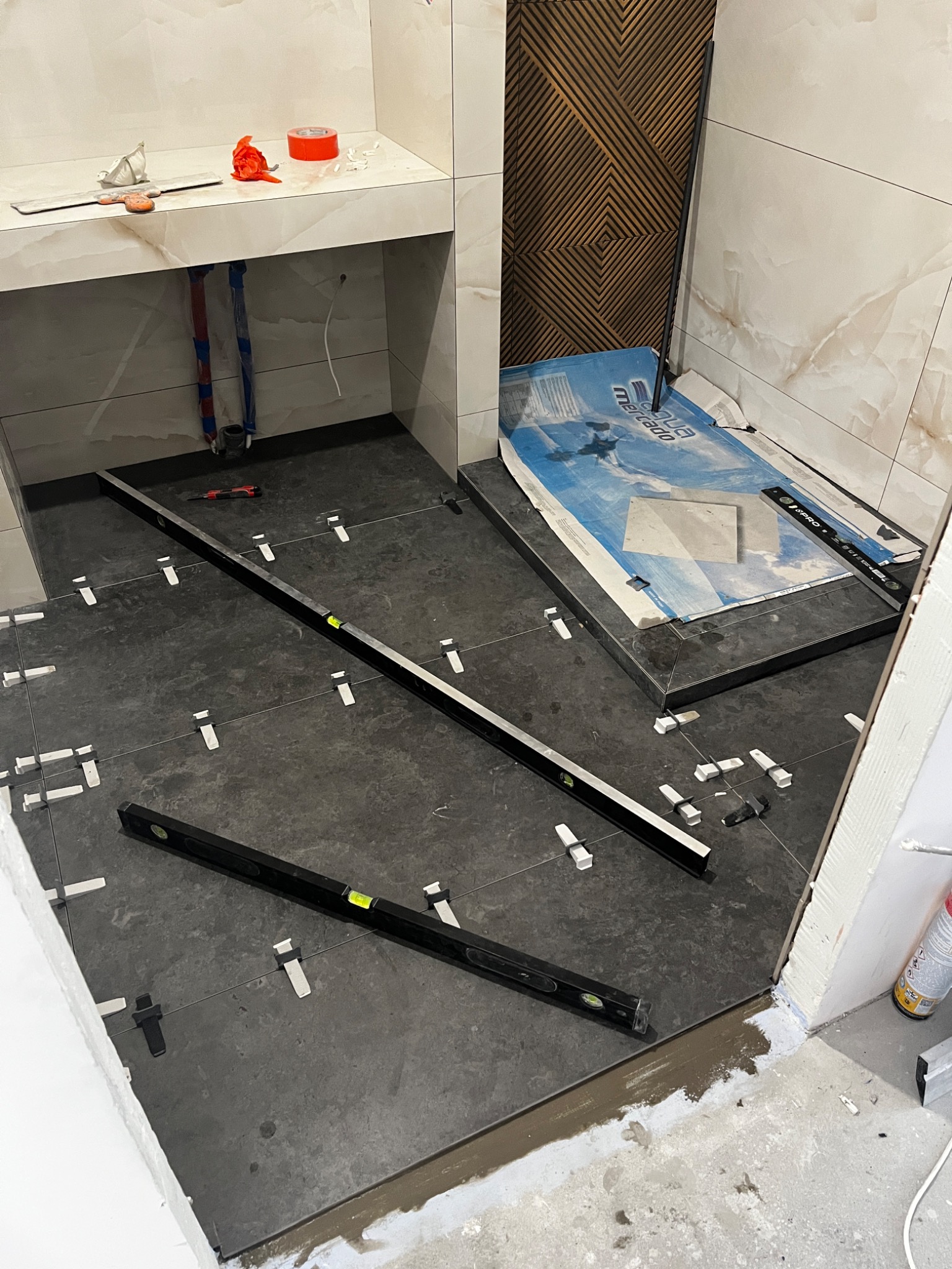 Układanie ciemnych płytek podłogowych w łazience z użyciem poziomicy i klipsów dystansowych, widoczne przygotowane rury i narzędzia.