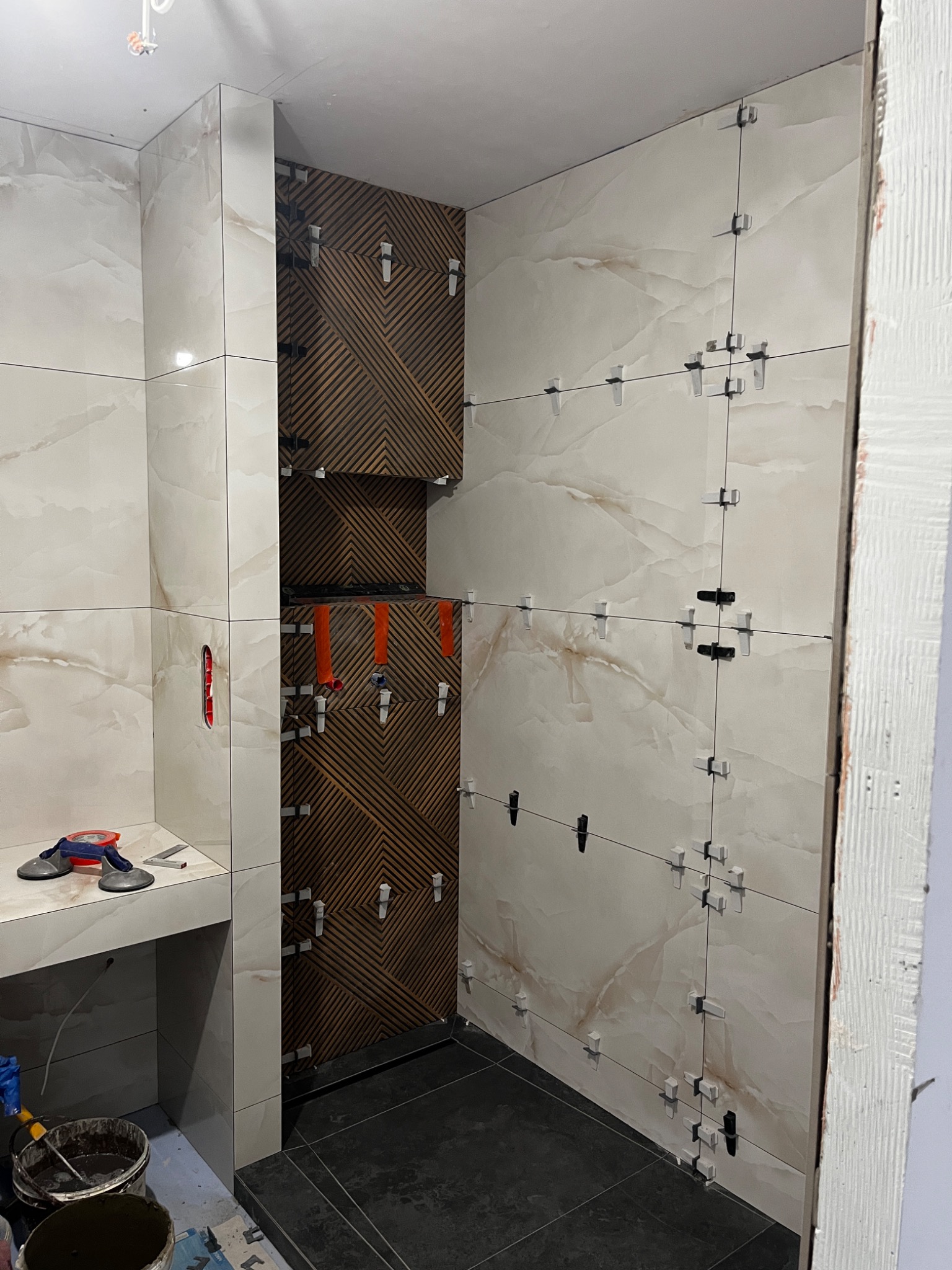 Ściany łazienki w trakcie układania płytek: beżowe płytki imitujące marmur z białymi krzyżykami dystansowymi sąsiadują z ciemnobrązowymi płytkami ułożonymi w geometryczny wzór; widoczne narzędzia...