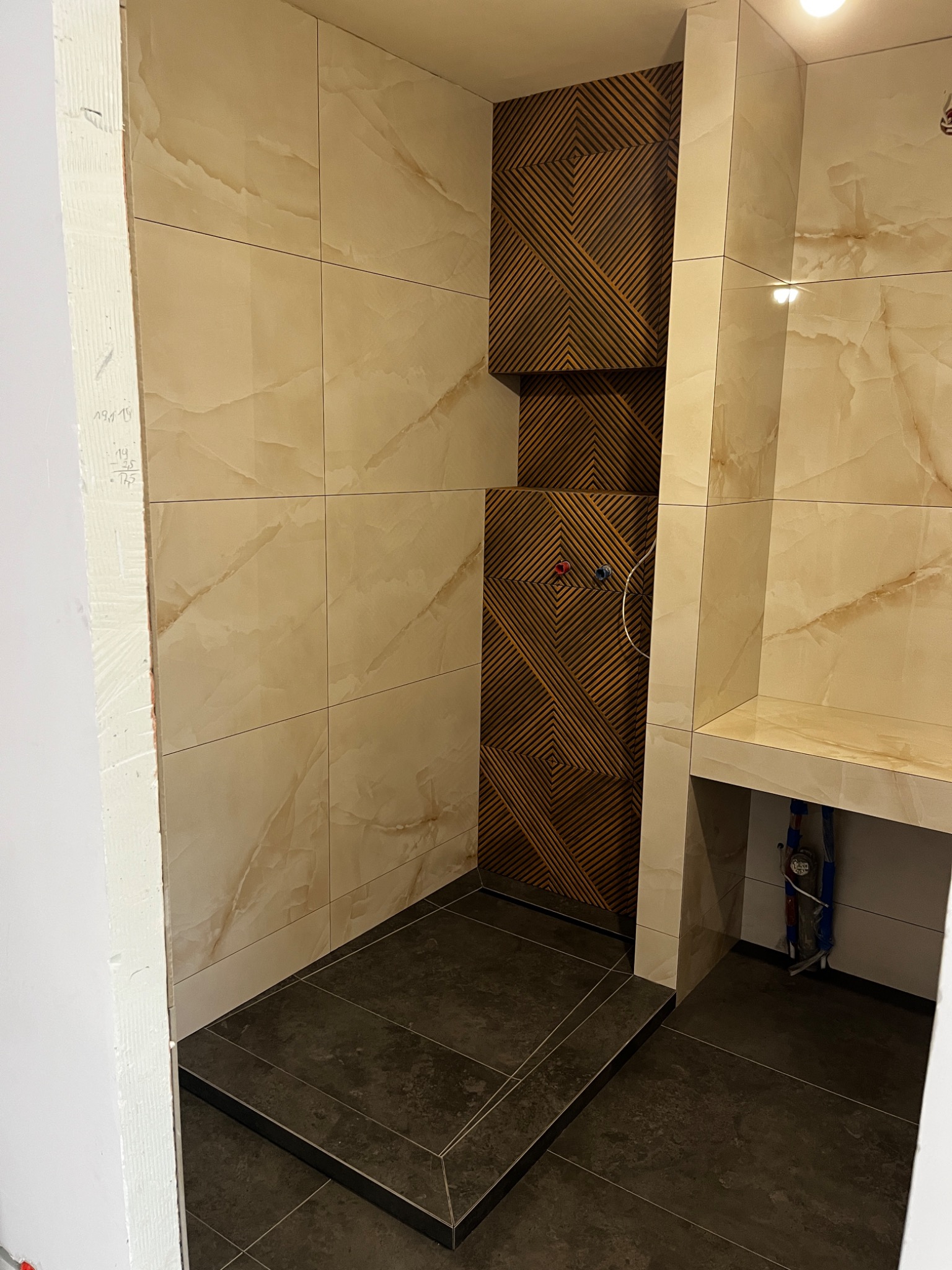 Płytki imitujące marmur na ścianach kabiny prysznicowej z dekoracyjną ścianką z drewnopodobnych paneli z geometrycznym wzorem; ciemnoszara podłoga z płytek.