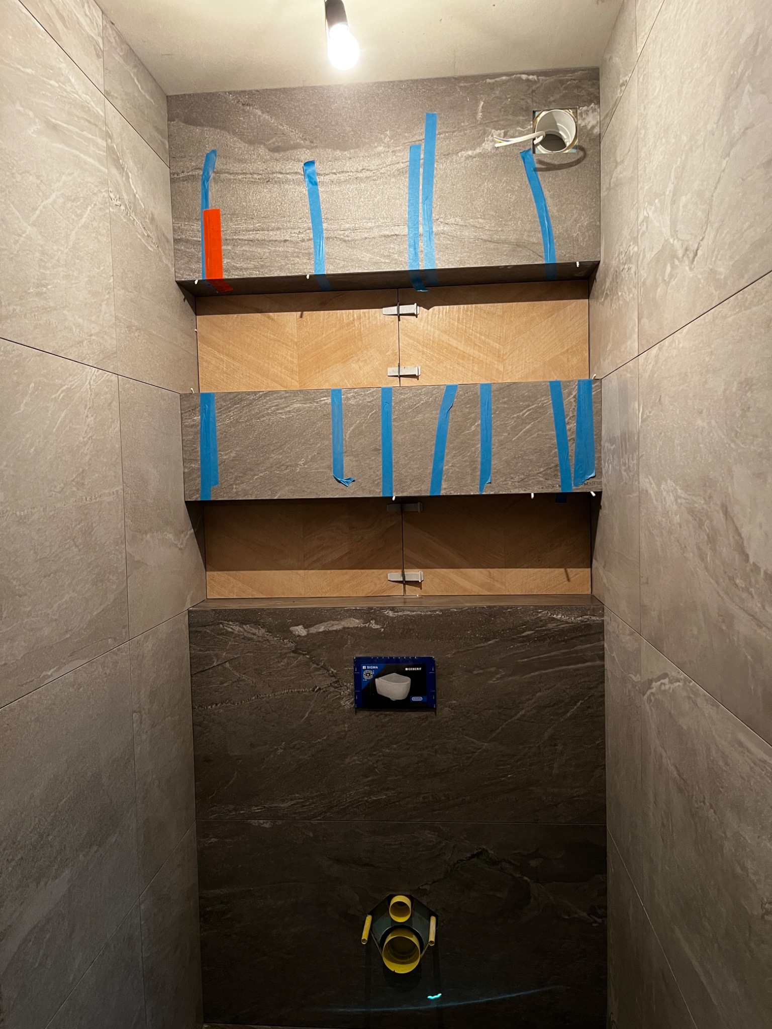 Ściana łazienki w trakcie układania płytek imitujących kamień, zabezpieczonych niebieską taśmą, widoczny stelaż podtynkowy WC i fragment instalacji wodnej