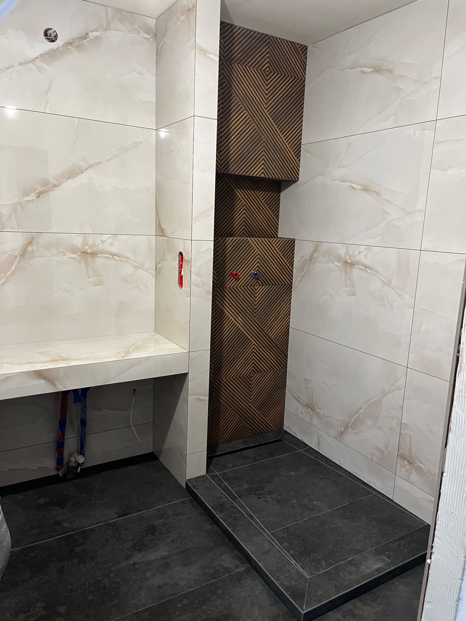 Nowoczesna łazienka w trakcie wykańczania: beżowe płytki imitujące marmur, ciemna podłoga i dekoracyjna ściana z drewnianych listew nad prysznicem. Widoczne instalacje wodne i fragment...