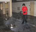 Czyszczenie posadzki w garażu podziemnym za pomocą profesjonalnej maszyny czyszczącej. Operator w czerwonej koszulce i czapce roboczej. Widoczna para wodna i brudna woda usuwana z powierzchni.
