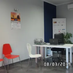 Biuro z trzema krzesłami, biurkiem z laptopem i kalendarzem na ścianie, plakat z logo firm finansowych i ubezpieczeniowych.