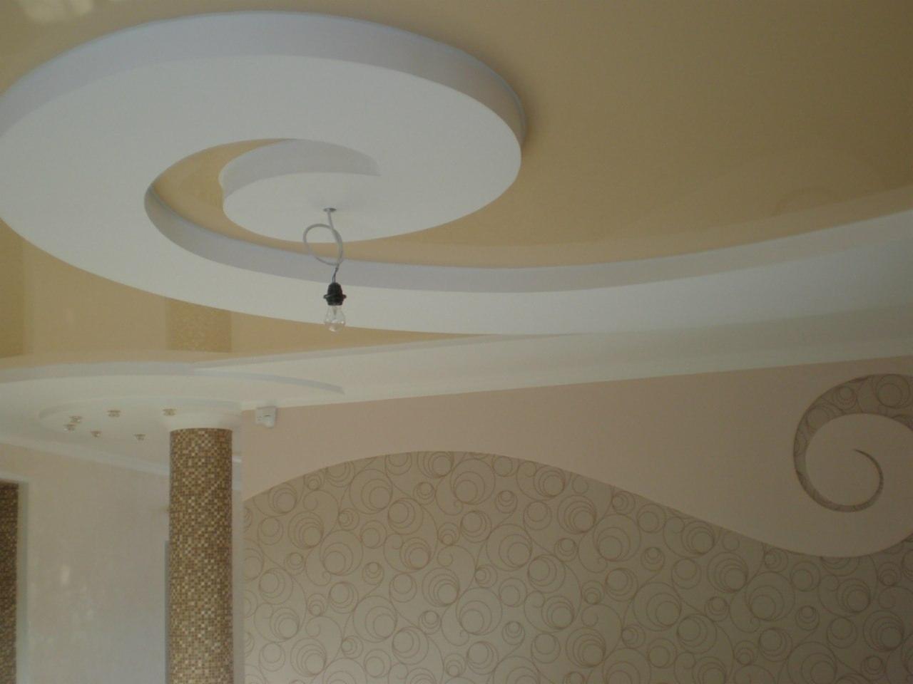 Kreatywny sufit podwieszany w kształcie spirali z dekoracyjnym oświetleniem i kolumną wykończoną mozaiką, na ścianie wzorzysta tapeta.