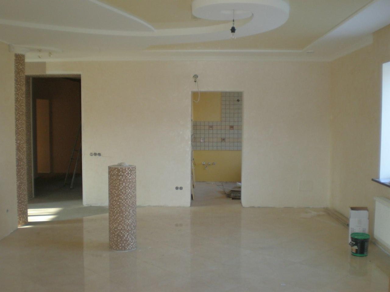 Wnętrze w trakcie wykańczania: położona jasna podłoga, ściany przygotowane pod malowanie, widoczny fragment dekoracyjnego sufitu i resztki materiałów budowlanych przy grzejniku.