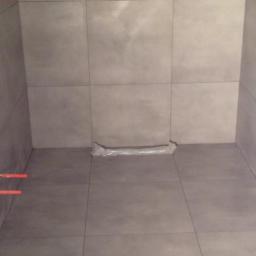 VP WORK&DESIGN sp. z.o.o. - Surowa, szara łazienka w trakcie wykańczania, widoczne rury i folia ochronna na podłodze, płytki imitujące beton na ścianach i podłodze.