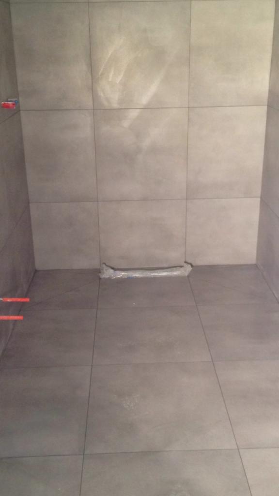 Surowa, szara łazienka w trakcie wykańczania, widoczne rury i folia ochronna na podłodze, płytki imitujące beton na ścianach i podłodze.