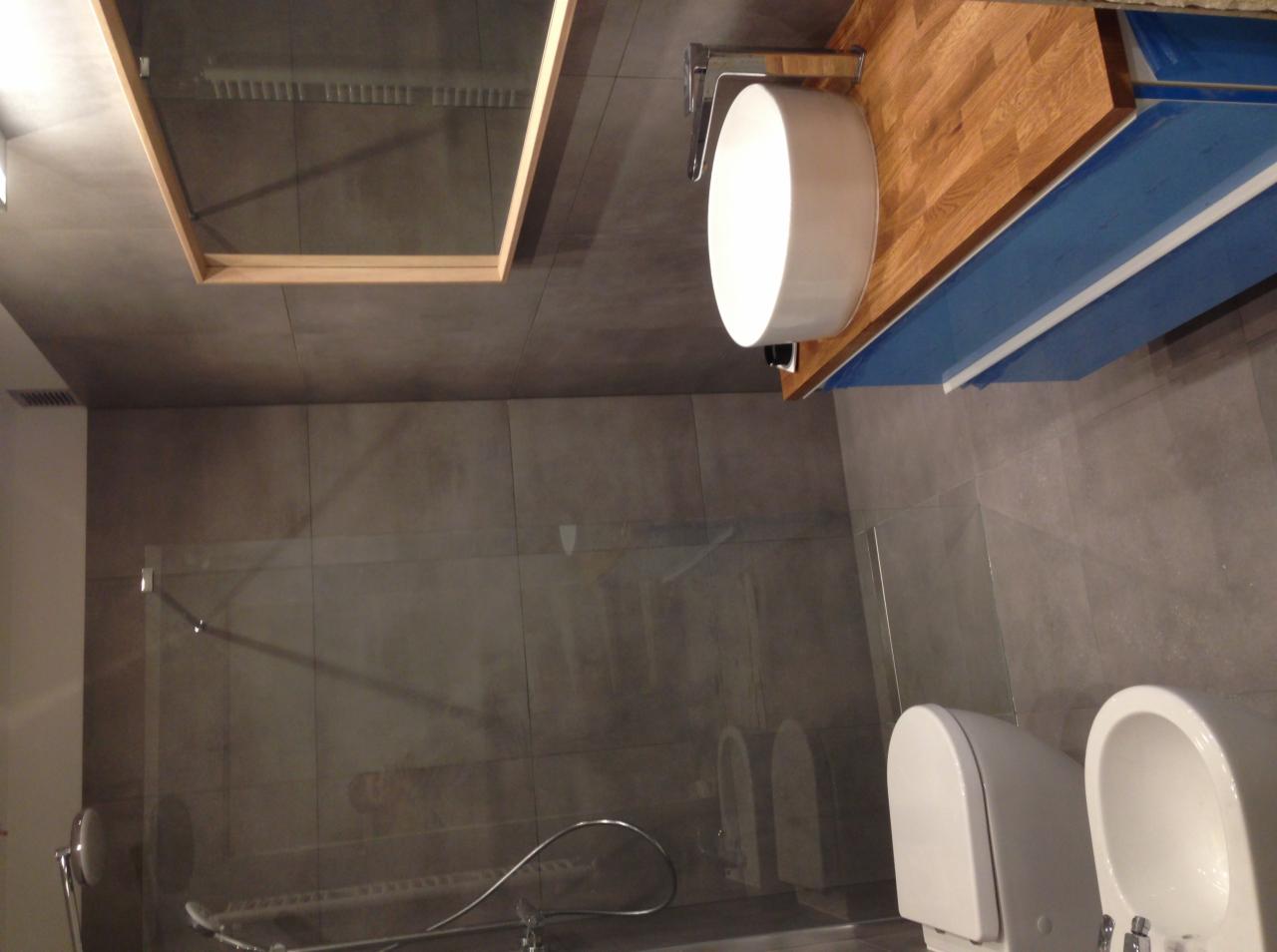 Nowoczesna łazienka z szarą, wielkoformatową płytką, drewnianym blatem pod umywalką nablatową, szklaną kabiną prysznicową i podwieszaną miską WC z bidetem.