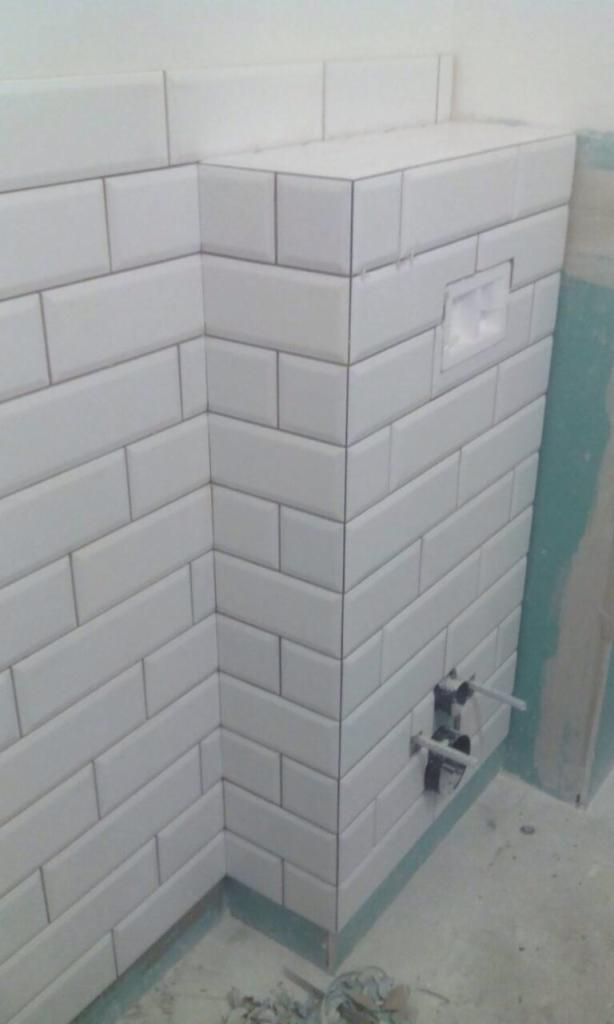 Świeżo ułożone białe, prostokątne płytki typu metro w rogu łazienki, widoczne instalacje wodne i fragment ściany przygotowanej do dalszych prac.