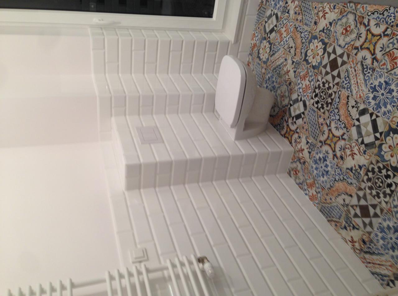 Biała łazienka wykończona podłużnymi płytkami, z ozdobną ścianą w kolorowe wzory i nowoczesnym grzejnikiem drabinkowym.