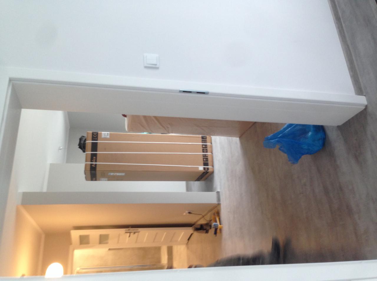 Widok przez otwarte drzwi na podłogę z panelami, na której leżą kartony z nowymi meblami i niebieska torba foliowa, widoczny fragment sufitu z włącznikiem światła.