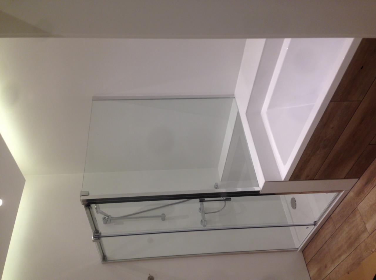 Nowoczesna kabina prysznicowa z przesuwnymi drzwiami szklanymi, chromowane wykończenia, widok z góry na białą obudowę wanny i fragment drewnianej podłogi.