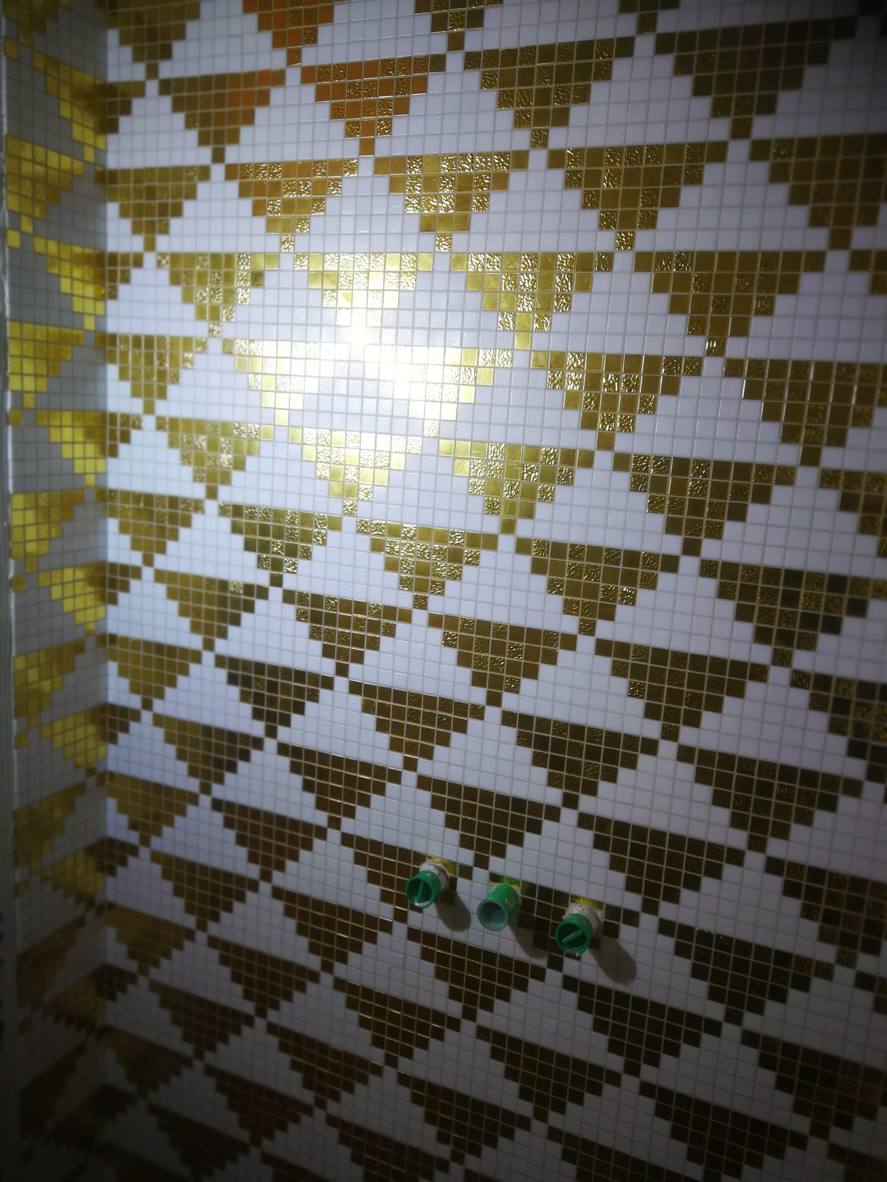 Ściana w łazience wyłożona białymi i złotymi mozaikami tworzącymi geometryczny wzór, z widocznymi trzema zielonymi rurami instalacji wodnej.