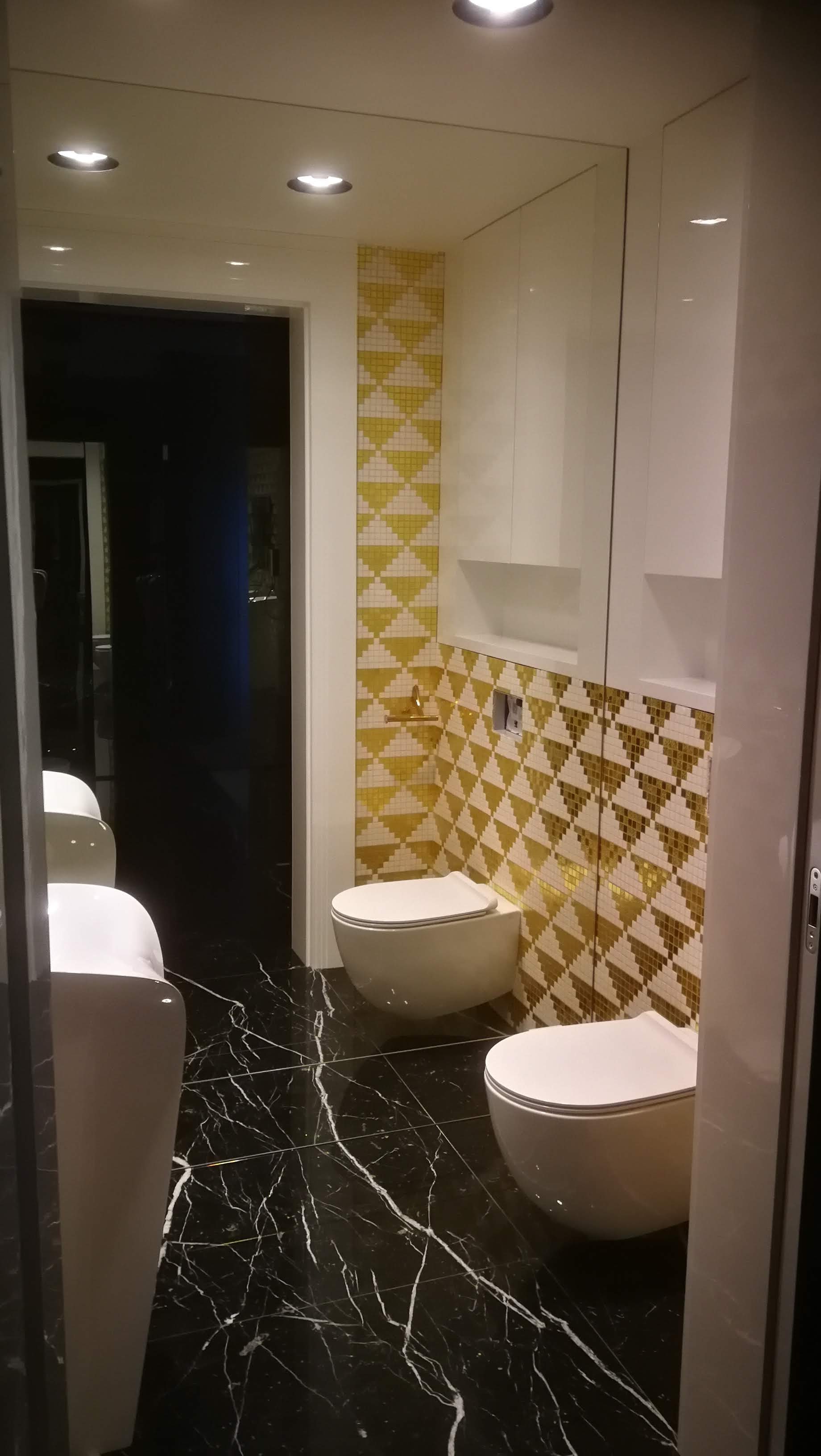 Nowoczesna łazienka z czarną marmurową podłogą, geometryczną mozaiką w odcieniach złota i bieli na ścianie oraz podwieszanymi toaletami.