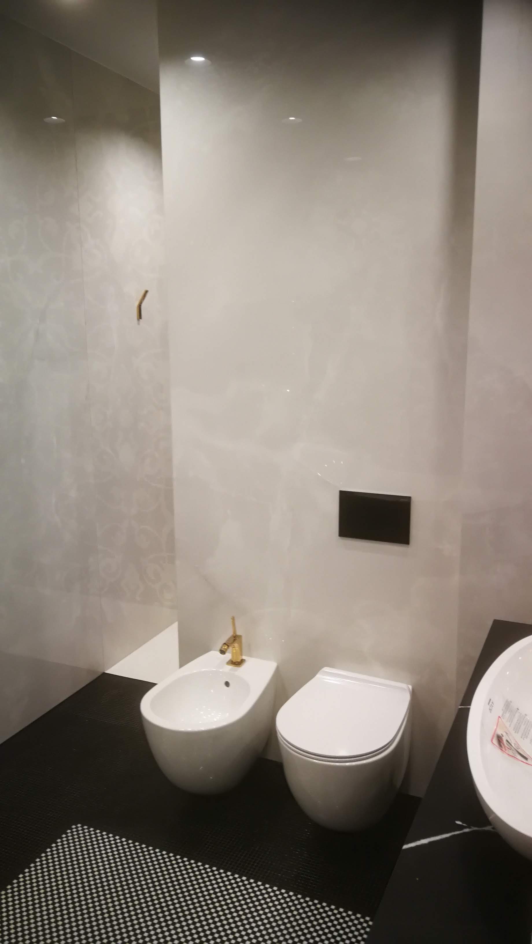 Nowoczesna łazienka w odcieniach bieli i czerni z armaturą w kolorze złota: wisząca toaleta, bidet z baterią i umywalka na blacie z czarnego marmuru, płytki podłogowe w czarno-białą szachownicę.