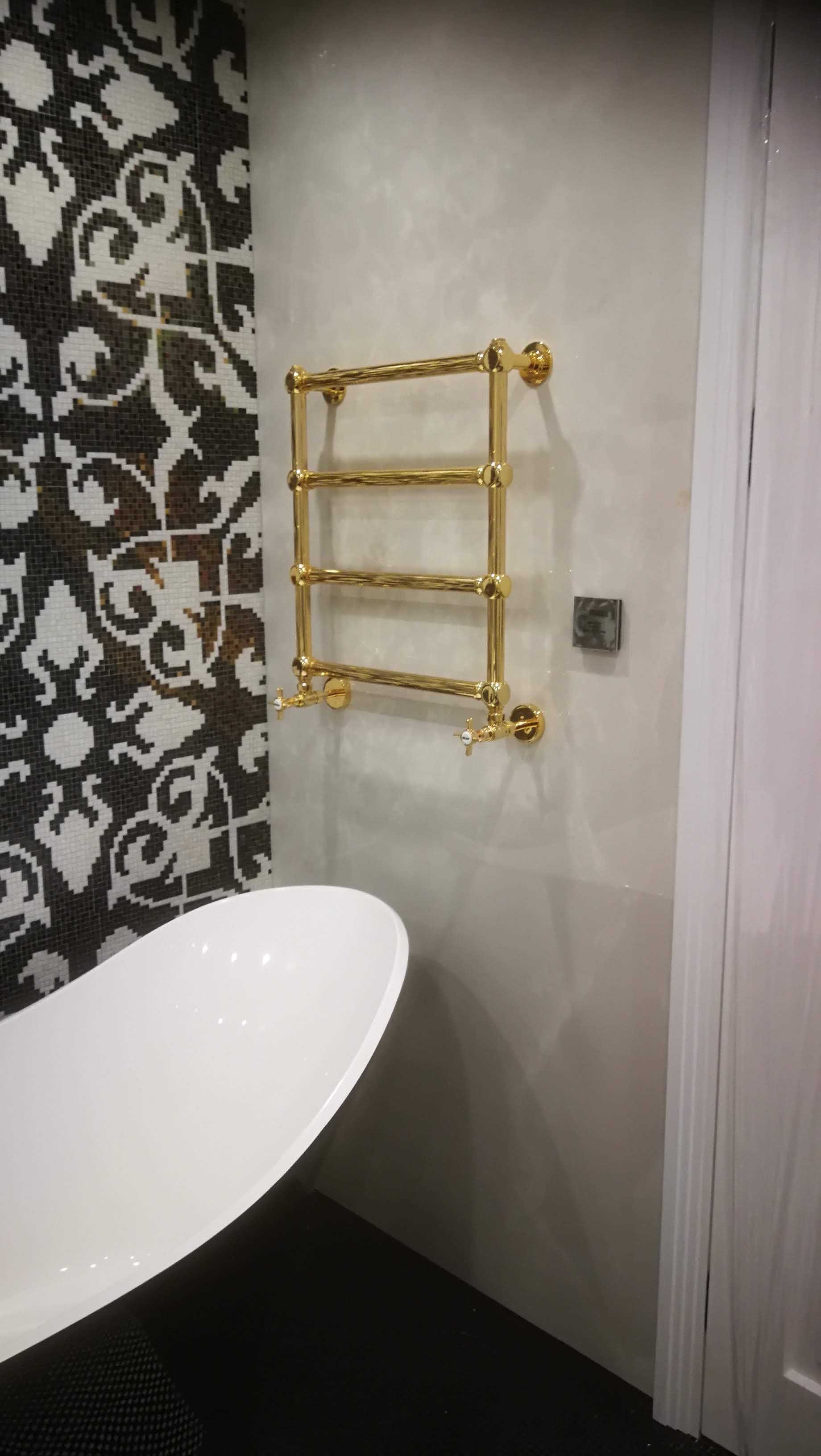 Wykończona łazienka z wolnostojącą białą wanną, dekoracyjną mozaiką na ścianie i złotym grzejnikiem drabinkowym.