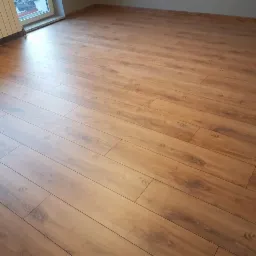 Ukończona podłoga z paneli laminowanych w odcieniach ciepłego brązu, ułożona wzdłuż okna z widocznym grzejnikiem żeberkowym.