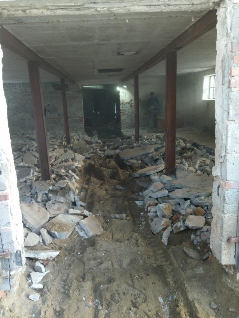 Wnętrze budynku w trakcie rozbiórki, widoczne stalowe słupy podtrzymujące strop, gruz i ziemia na podłodze, ślady opon pojazdu budowlanego, w tle sylwetka osoby.