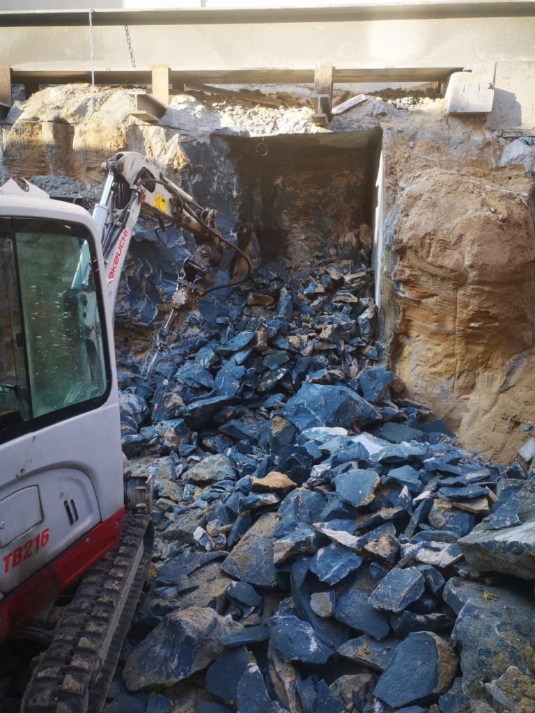 Mini koparka Takeuchi TB216 z hydraulicznym młotem wykuwa otwór w ziemi, otoczony stertą ciemnych odłamków skalnych. Zabezpieczenie stalowymi belkami nad wykopem.