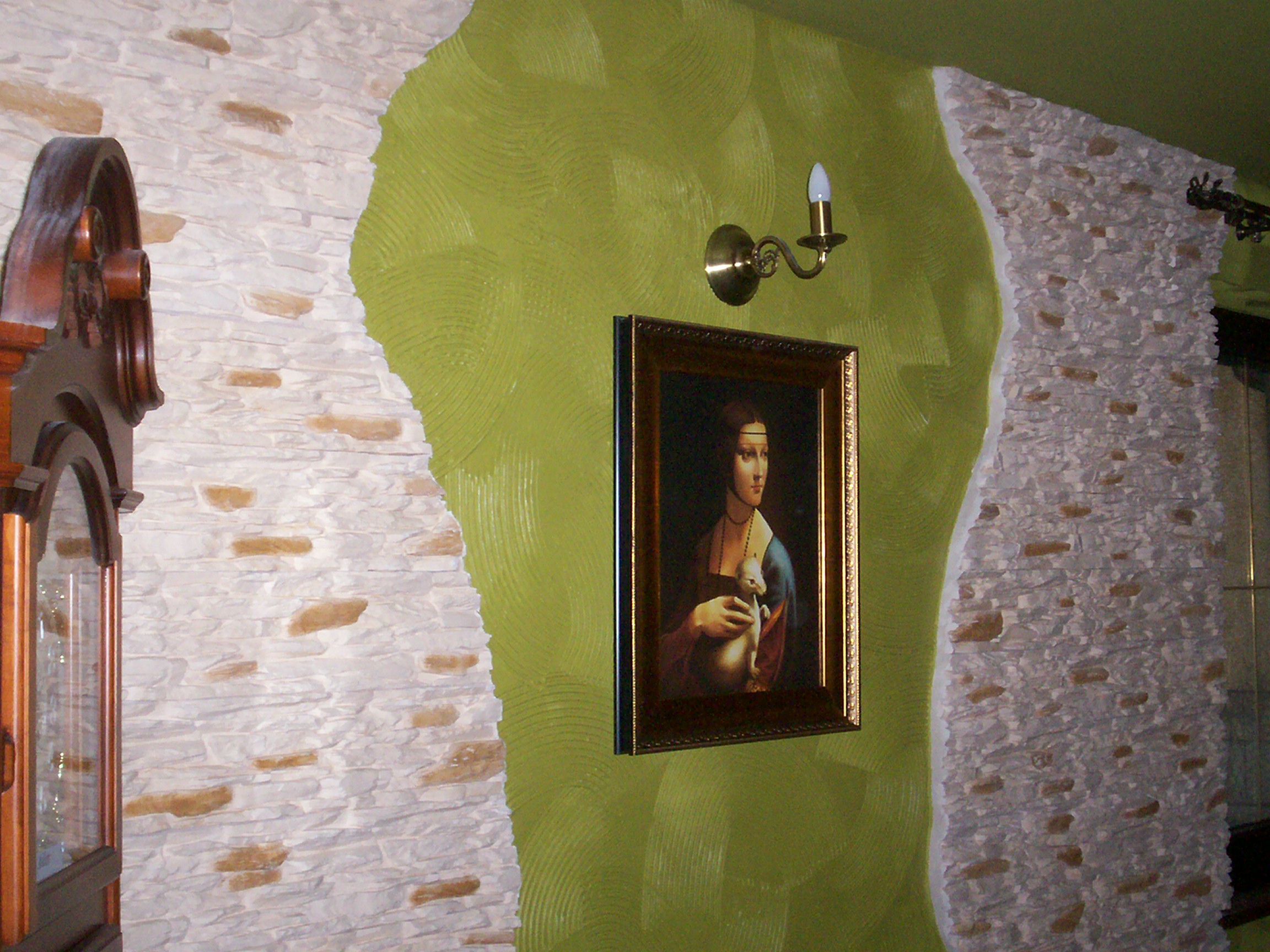 Wykończenie wnętrza z dekoracyjną ścianą z kamienia, zieloną ścianą z efektem okręgów i obrazem w złotej ramie pod kinkietem.