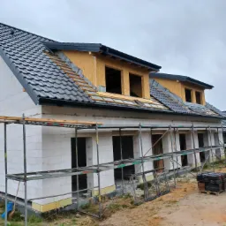 Budowa szeregowca z białego bloczka, częściowo pokrytego czarną dachówką, z widocznym oknem dachowym w trakcie montażu i rusztowaniem.