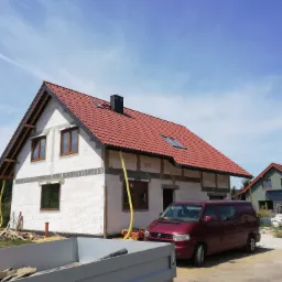 Niedokończony dom murowany z czerwonym dachem, widoczny fragment przyczepy i bordowy van na tle błękitnego nieba.
