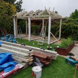 Budowa drewnianej altany w ogrodzie: widoczne elementy konstrukcyjne, narzędzia, materiały budowlane, drabina, worki z odpadami i skrzynia z roślinami w tle.