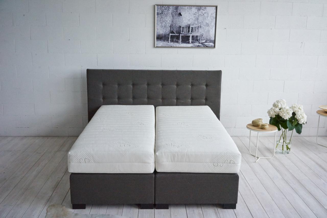 Dwa białe materace z szarym zagłówkiem na tle białej ściany z obrazem i stolikiem z kwiatami.