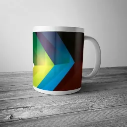 Biały kubek z abstrakcyjnym, geometrycznym wzorem z przewagą niebieskiego, zielonego, żółtego i brązowego, stojący na szarym, drewnianym blacie na tle jasnej ściany.