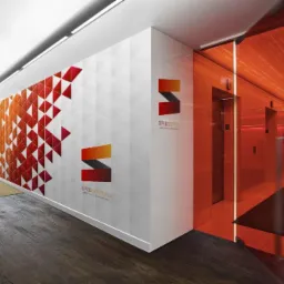 Wnętrze biura z geometrycznym wzorem w odcieniach czerwieni i pomarańczy na ścianie, logo firmy Spedsoft.eu i windami za szklaną, pomarańczową ścianą.