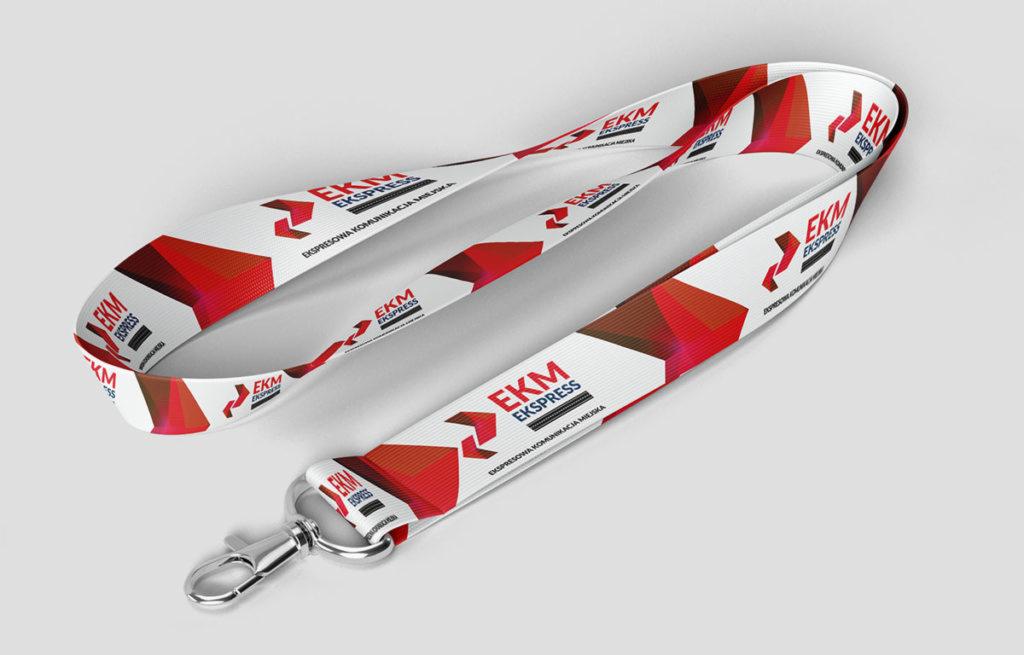 Biała smycz z powtarzającym się logo EKM Ekspress w czerwono-białej kolorystyce, leżąca na szarym tle, z metalowym karabińczykiem.