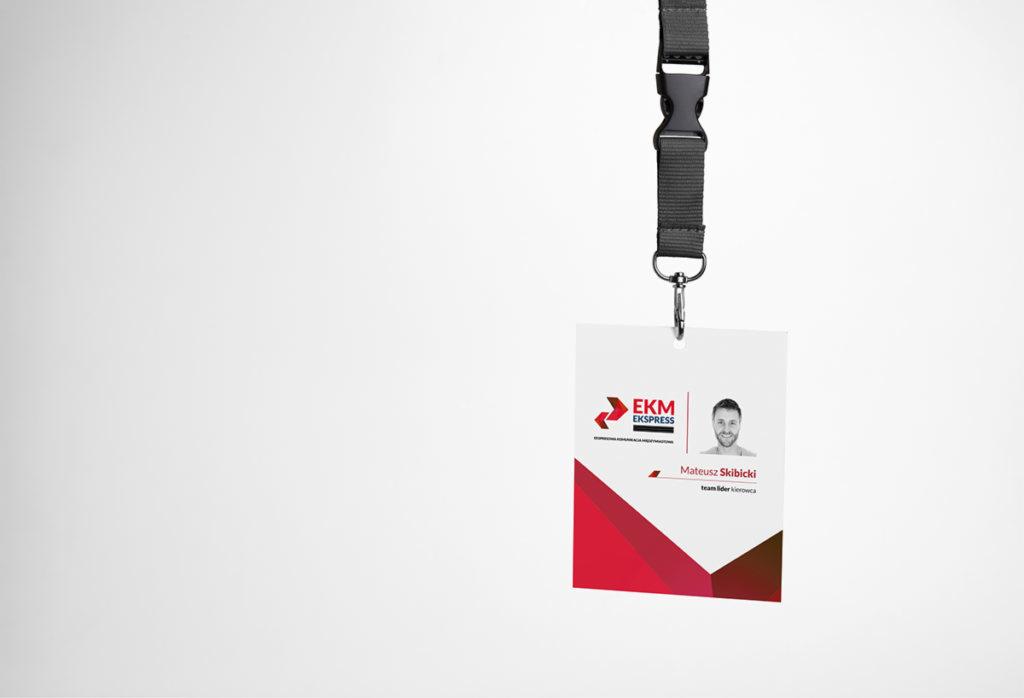 Identyfikator z logo firmy EKM Ekspress, zdjęciem pracownika Mateusza Skibickiego i informacją o stanowisku Team Lider Kierowca zawieszony na czarnej smyczy z klamrą.