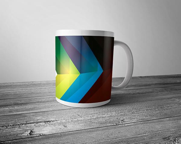 Biały kubek z abstrakcyjnym, geometrycznym wzorem z przewagą niebieskiego, zielonego, żółtego i brązowego, stojący na szarym, drewnianym blacie na tle jasnej ściany.