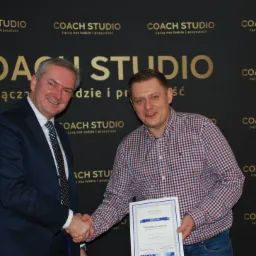 Dw&oacute;ch mężczyzn w uścisku dłoni na tle ściany z logo Coach Studio, jeden w garniturze, drugi w koszuli w kratę, trzyma dyplom z napisem 'Pan Andrzej Łukasiak'.