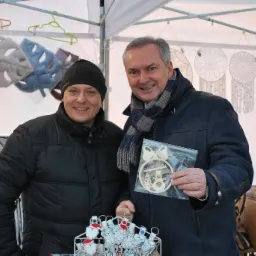 Dwóch uśmiechniętych mężczyzn na stoisku targowym prezentuje ręcznie robione ozdoby świąteczne, w tym drewnianą bombkę z motywem bałwana i choinek oraz zawieszki z postaciami Mikołaja i jednorożców.