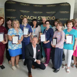 Grupa osób, w tym mężczyzna w garniturze, prezentuje certyfikaty ukończenia szkolenia Coach Studio na tle baneru z logo Coach Studio.