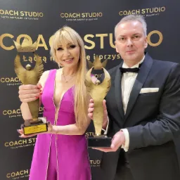 Uśmiechnięta kobieta w fuksjowej sukience i mężczyzna w smokingu trzymają złote statuetki z koroną na tle ściany z logo Coach Studio.