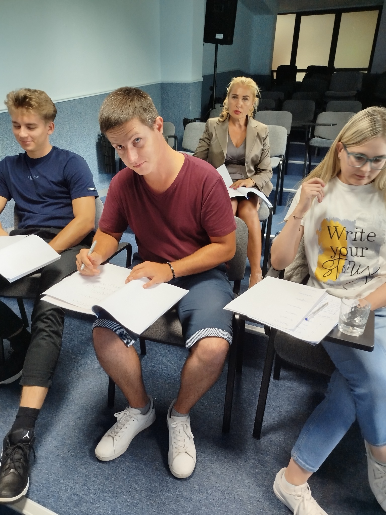 Uczestnicy szkolenia w Wojkowicach, siedzący przy stolikach i robiący notatki. W tle kobieta w żakiecie. Sala konferencyjna z niebieską wykładziną.