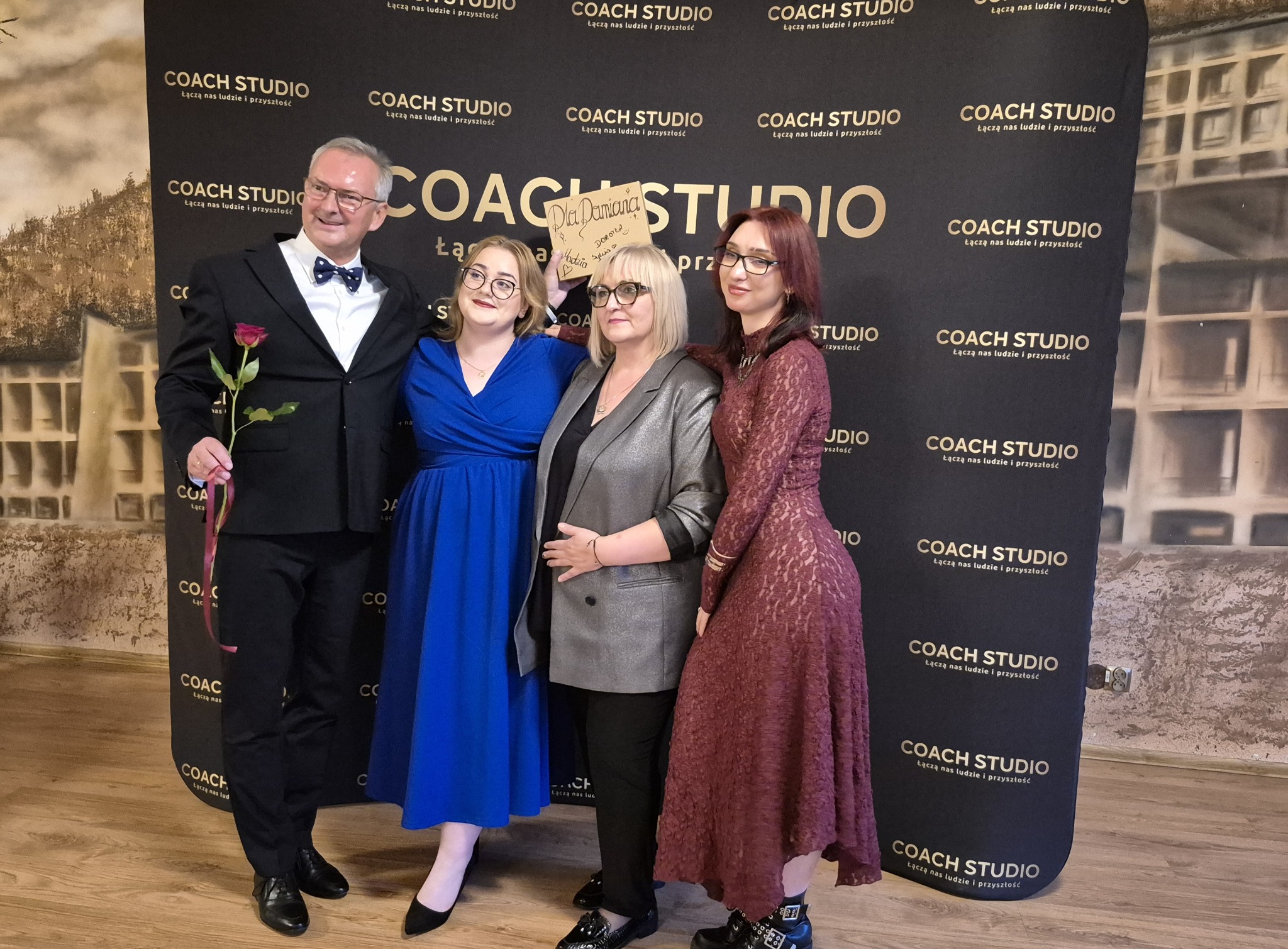 Grupa osób, w tym mężczyzna z różą, pozuje na tle ścianki z logo 'Coach Studio'. Jedna z kobiet trzyma kartkę z dedykacją. Uśmiechy, odświętne stroje.