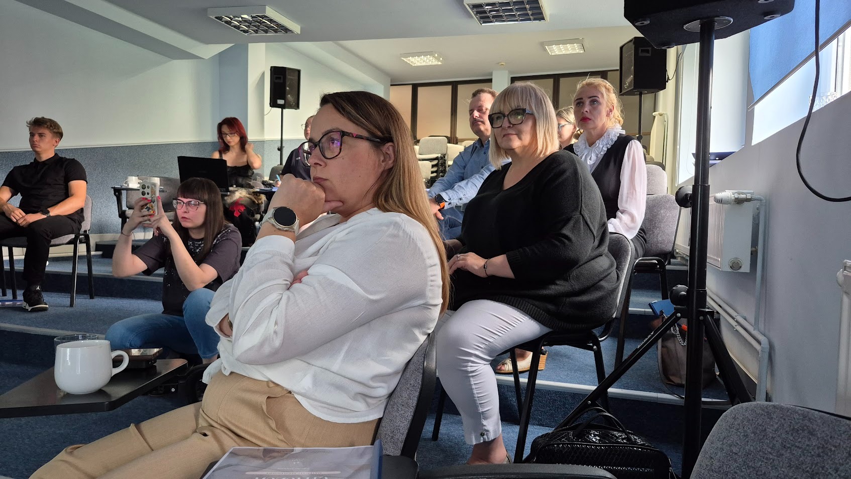 Uczestnicy w skupieniu podczas szkolenia w sali konferencyjnej, kobieta w białej koszuli z zegarkiem na pierwszym planie, osoby w tle siedzą na krzesłach.