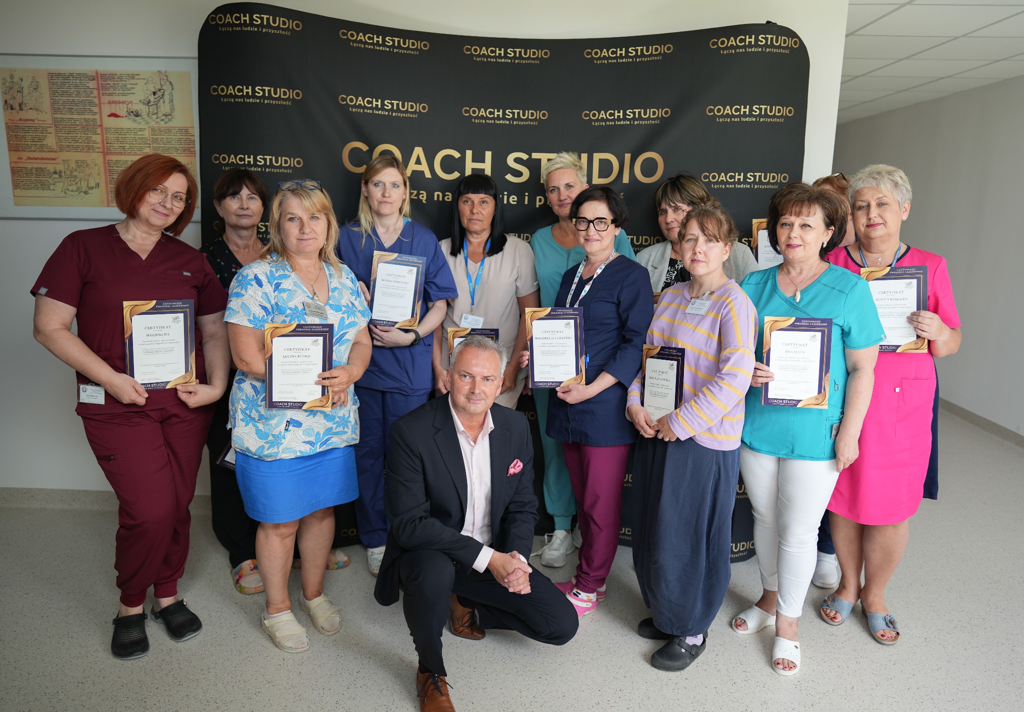 Grupa osób, w tym mężczyzna w garniturze, prezentuje certyfikaty ukończenia szkolenia Coach Studio na tle baneru z logo Coach Studio.