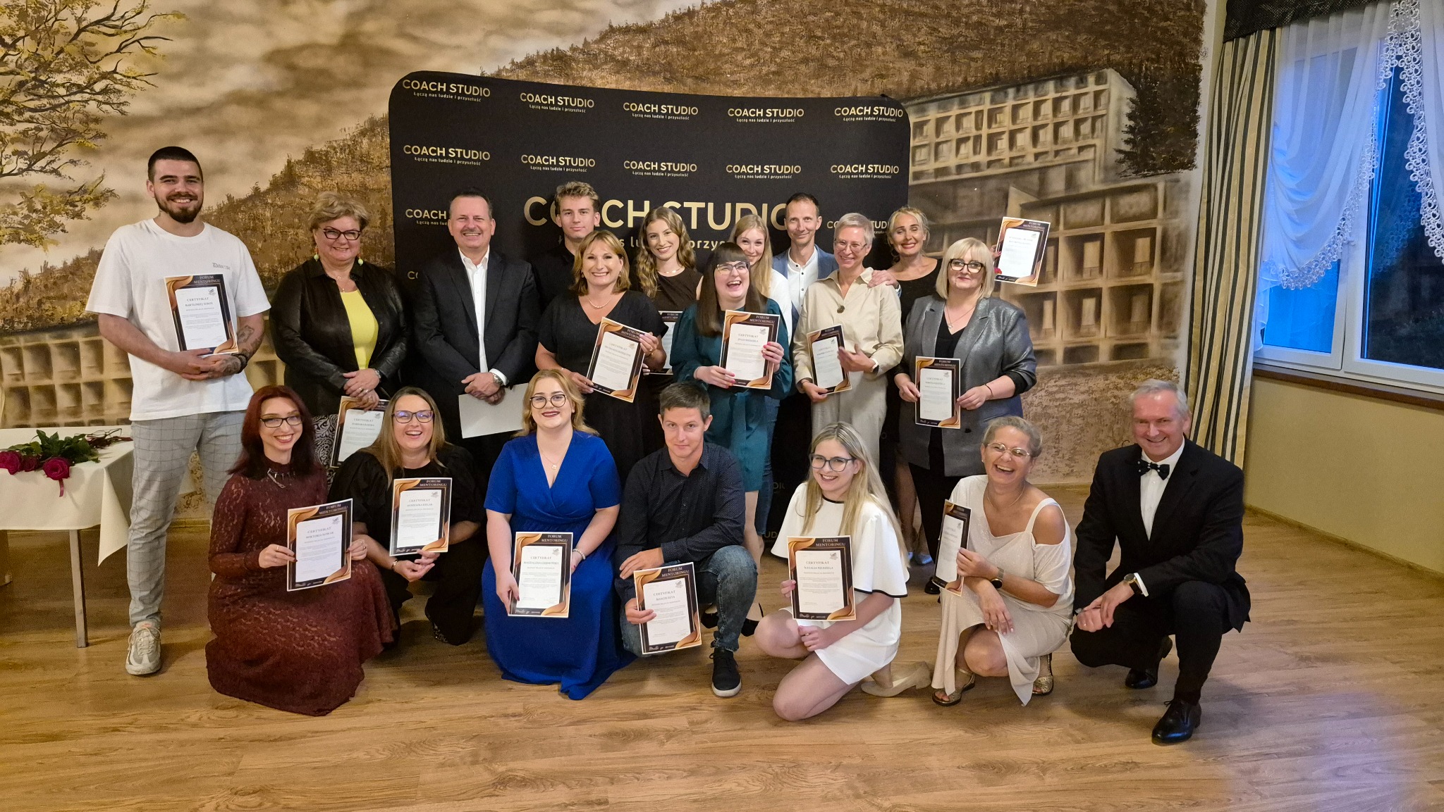 Grupa osób trzyma certyfikaty na tle ściany z logo Coach Studio. Uczestnicy szkolenia z uśmiechami prezentują dyplomy ukończenia kursu. W tle dekoracja imitująca budynek.