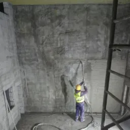 Pracownik w kasku i kamizelce odblaskowej aplikuje zaprawę na ścianę w surowym, betonowym pomieszczeniu za pomocą węża pod ciśnieniem, widoczna drabina i okno z kratami.