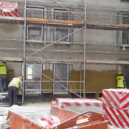Prace na rusztowaniu: montaż izolacji termicznej na elewacji budynku, widoczni pracownicy w żółtych kamizelkach, paczki materiału izolacyjnego Paroc na pierwszym planie.