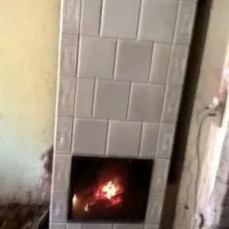 Wykończony piec kaflowy z palącym się ogniem w palenisku, obudowany beżowymi płytkami z dekoracyjnym obramowaniem, widoczne ślady prac remontowych na podłodze.