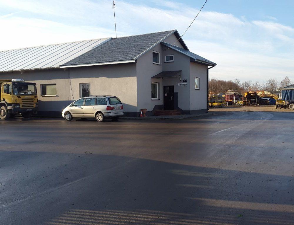 Żółta ciężarówka Scania i srebrny van zaparkowane przed budynkiem betoniarni z szarymi ścianami i dwuspadowym dachem; w tle składowane maszyny budowlane.