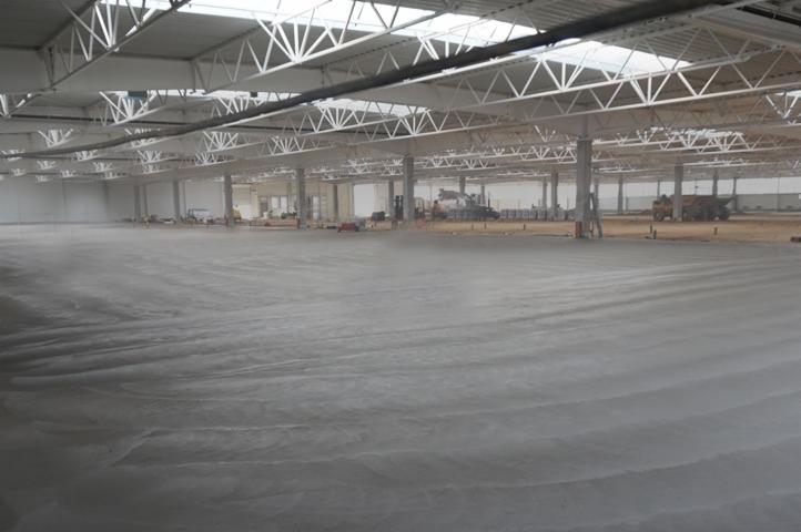 Wylany betonowy posadzka przemysłowa w hali magazynowej z widocznymi maszynami budowlanymi w tle, oświetlona światłem dziennym przez świetliki dachowe.